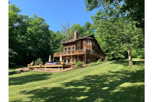 A large wooden house surrounded by lush greenery and a well-maintained lawn, featuring a deck and outdoor seating area.