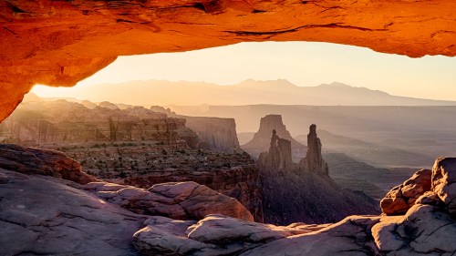 Sunset view through a rock arch, showcasing rugged cliffs and distant mountains in a serene landscape.