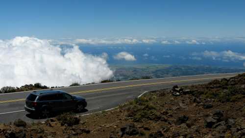 A car parked on a winding road overlooking clouds and a coastal landscape below.