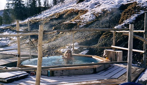 A person relaxes in a hot spring surrounded by snow-covered ground and wooden structures. Steam rises in the cold air.