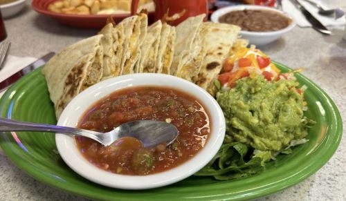 A plate of quesadillas with salsa, guacamole, and a side salad on a green dish, with additional food in the background.