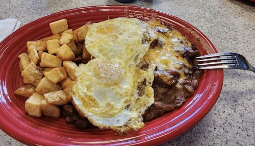 A red plate with two fried eggs, cheese-covered beans, and golden-brown potato cubes.