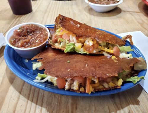 A blue plate with two crispy tacos filled with lettuce, tomatoes, cheese, and chicken, served with a side of salsa.