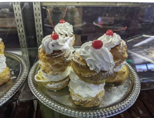 A silver tray holds stacked cream puffs topped with whipped cream and cherries, displayed in a glass case.
