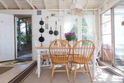 Cozy dining area with wooden chairs, a table, and hanging cookware, surrounded by natural light and greenery.