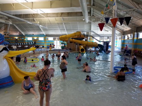 A busy indoor pool with a water slide, children playing, and adults relaxing in the water. Colorful decorations are visible.