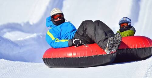Two people in winter gear sit on red snow tubes, sliding down a snowy slope. Bright blue sky in the background.