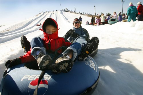 A child and an adult sit in a snow tube at a snowy hill, ready to slide down with others in the background.