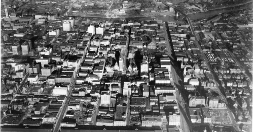 Aerial view of a cityscape featuring numerous buildings, streets, and a river in the background. Black and white image.