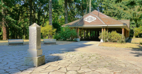 A stone monument stands in front of a covered pavilion surrounded by trees in a park setting.