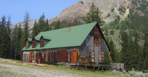 An old, weathered wooden building with a green roof, surrounded by trees and mountains under a clear blue sky.