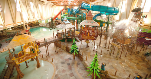 A vibrant indoor water park featuring slides, play structures, and a large pool area with trees and water features.
