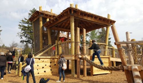 A wooden playground structure with slides and climbing areas, surrounded by children and adults enjoying the outdoors.