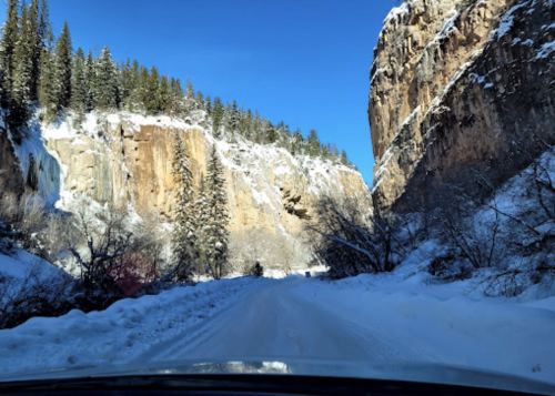 Snow-covered road winding through tall cliffs and pine trees under a clear blue sky.