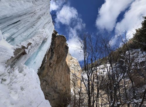 A snowy landscape with icy cliffs under a blue sky and scattered clouds, surrounded by bare trees.