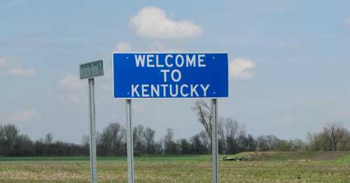 A blue welcome sign for Kentucky stands beside a road, with a clear sky and trees in the background.