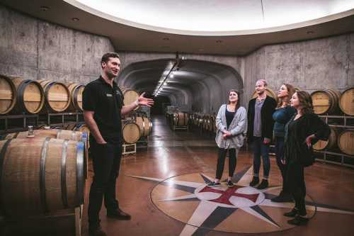 A guide speaks to a group of four people in a wine cellar surrounded by wooden barrels.