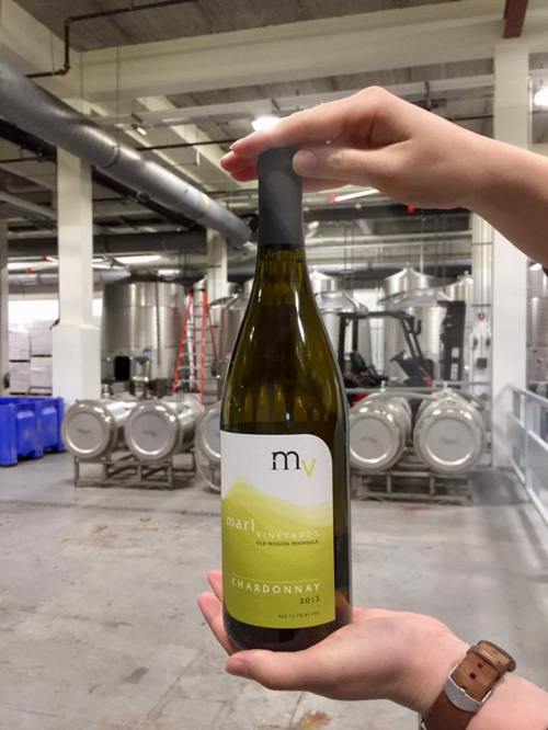 A hand holds a bottle of Chardonnay in a winery, with stainless steel tanks and barrels in the background.
