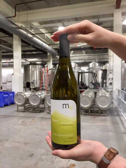 A hand holds a bottle of Chardonnay in a winery, with stainless steel tanks and barrels in the background.