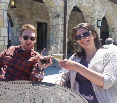 A man and woman clink their wine glasses together, smiling, seated at an outdoor table with a stone building in the background.