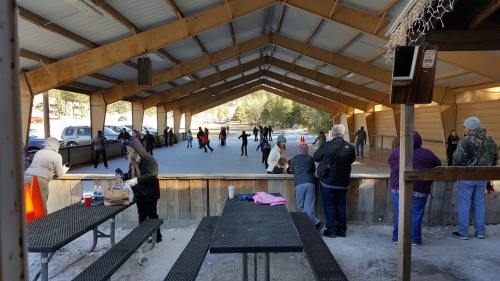 A covered ice skating rink with people skating and socializing, surrounded by trees and parked cars in the background.