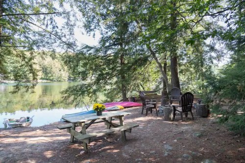 A serene lakeside scene with a picnic table, chairs, and a kayak, surrounded by trees and calm water.