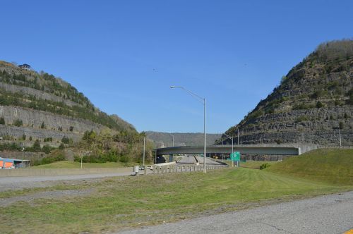 A highway curves between steep, rocky hills under a clear blue sky. Green grass lines the roadside.