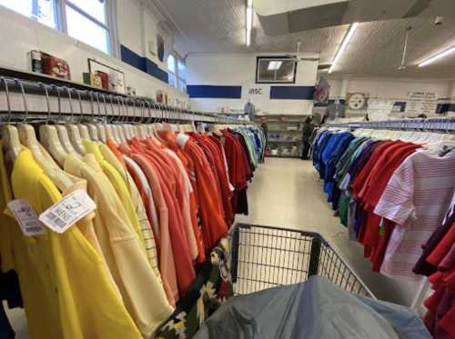 A clothing store aisle filled with colorful shirts on hangers, with a shopping cart in the foreground.