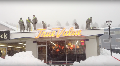 Workers in winter gear clear snow from the roof of a True Value store, surrounded by heavy snowfall.