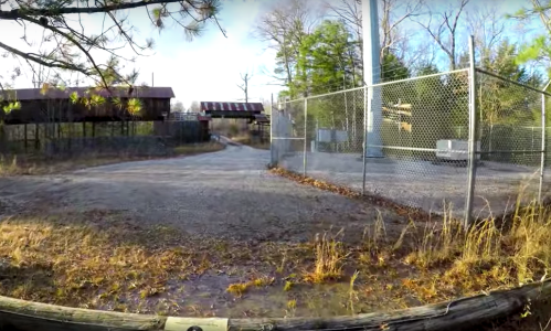 A gravel path leads through a fenced area with trees and abandoned buildings in the background.