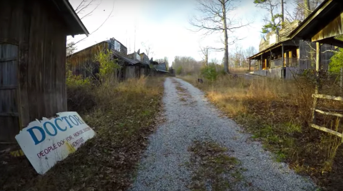 A gravel path leads through an overgrown area with abandoned buildings and a weathered sign reading "DOCTOR."