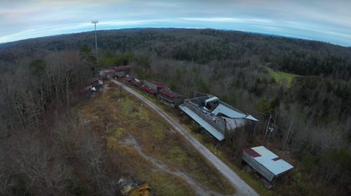 Aerial view of an abandoned site with dilapidated buildings surrounded by dense forest and a cell tower in the distance.