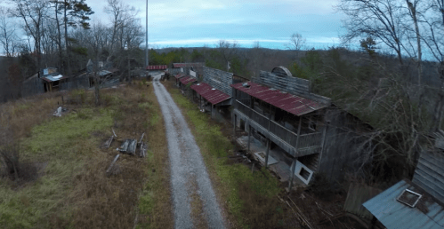 Aerial view of an abandoned building complex along a gravel path, surrounded by overgrown grass and trees.