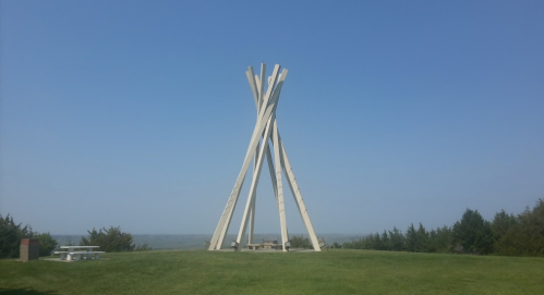 A tall, abstract sculpture made of wooden beams stands on a grassy hill under a clear blue sky.