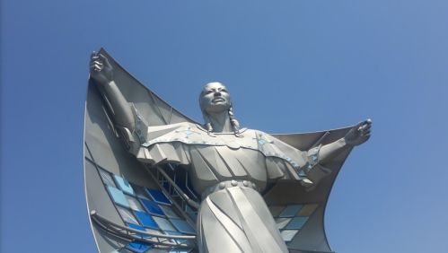 A large metallic statue of a woman with outstretched arms, set against a clear blue sky.