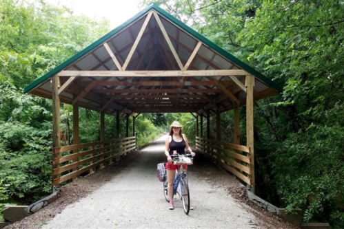A woman rides a bicycle under a wooden covered bridge surrounded by lush greenery.