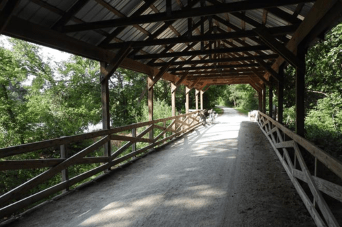 A covered wooden bridge with a dirt path, surrounded by lush greenery and trees.