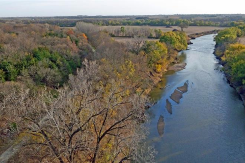 Aerial view of a winding river surrounded by trees in autumn colors and open fields.