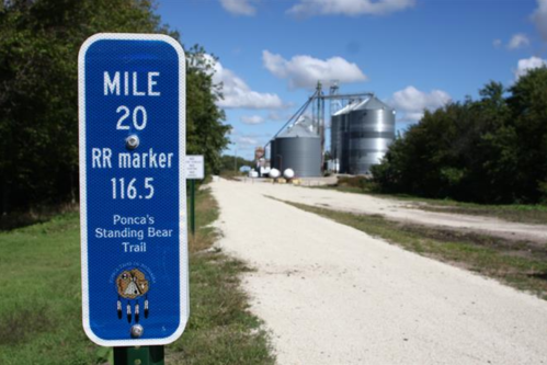 Blue railroad marker indicating Mile 20, with silos and a dirt path in the background under a partly cloudy sky.