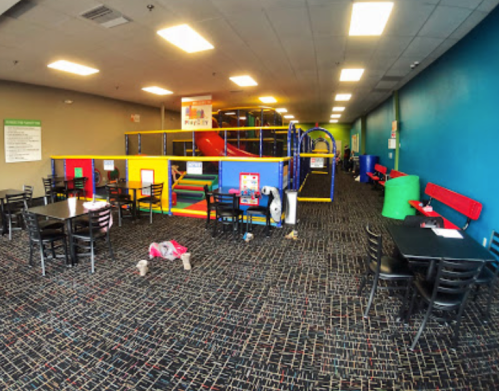 A colorful indoor play area with climbing structures, tables, and chairs, featuring a child playing on the floor.