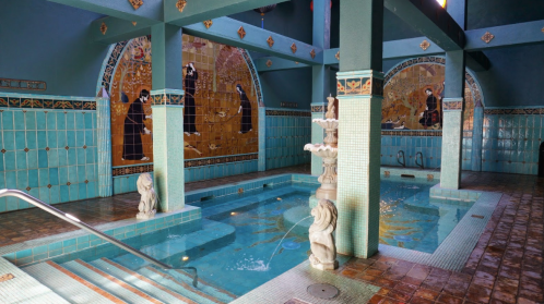 A serene indoor pool area with turquoise tiles, decorative murals, and a fountain surrounded by elegant architecture.