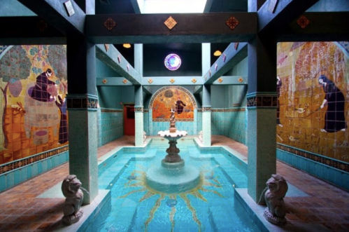 A serene indoor pool area with ornate tile work, a central fountain, and colorful murals on the walls.
