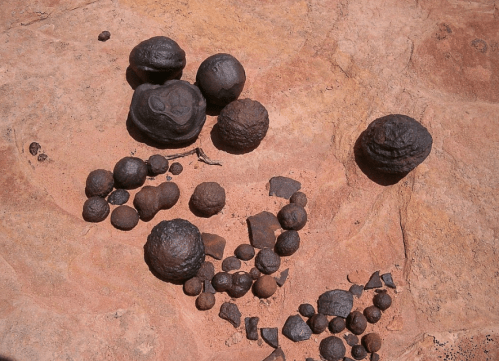 A collection of dark, round stones and fragments scattered on a reddish-brown rocky surface.