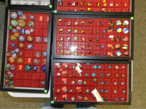 A collection of colorful marbles displayed in trays with red felt lining, organized in a grid pattern.
