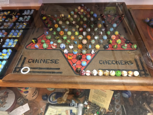 A vintage Chinese checkers board with colorful marbles, displayed in a glass case alongside various marbles and collectibles.