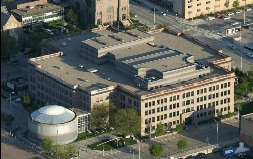 Aerial view of a large, modern building with a circular structure nearby, surrounded by trees and city streets.