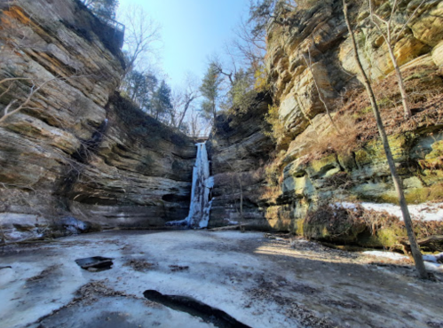 A frozen waterfall cascades down rocky cliffs, surrounded by trees and icy ground in a serene winter landscape.
