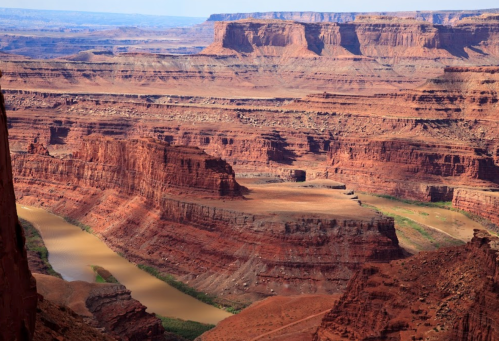 A vast canyon landscape with red rock formations and a winding river below, under a clear blue sky.