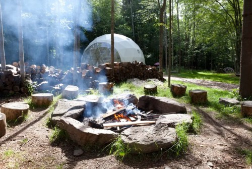 A campfire surrounded by logs, with smoke rising, and a geodesic dome in a wooded area.