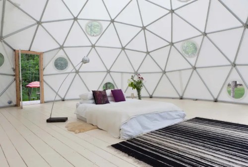 Interior of a geodesic dome with a bed, rug, and natural light from circular windows. A door leads outside.